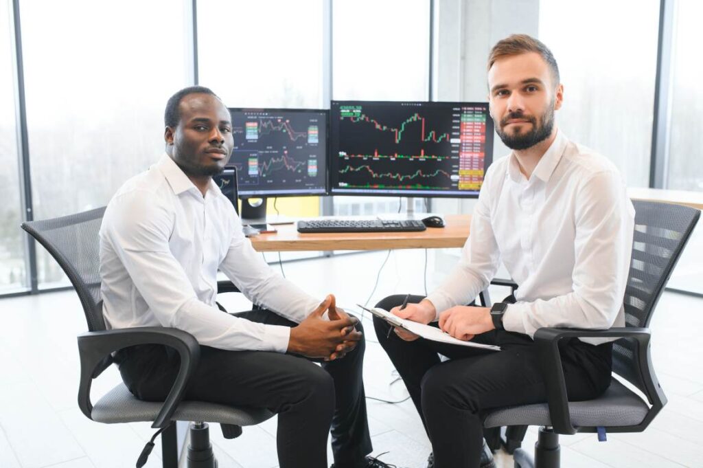 Two diverse colleagues traders talking to each other, sitting in the office